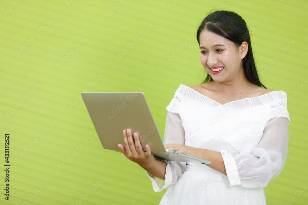 Asian smiling plump woman wearing white dress holding and looking at laptop computer. Lady asia standing is happy to working in green background. Asian smiling plump woman wearing white dress holding and looking at laptop computer. Lady asia standing is happy to working in green background.