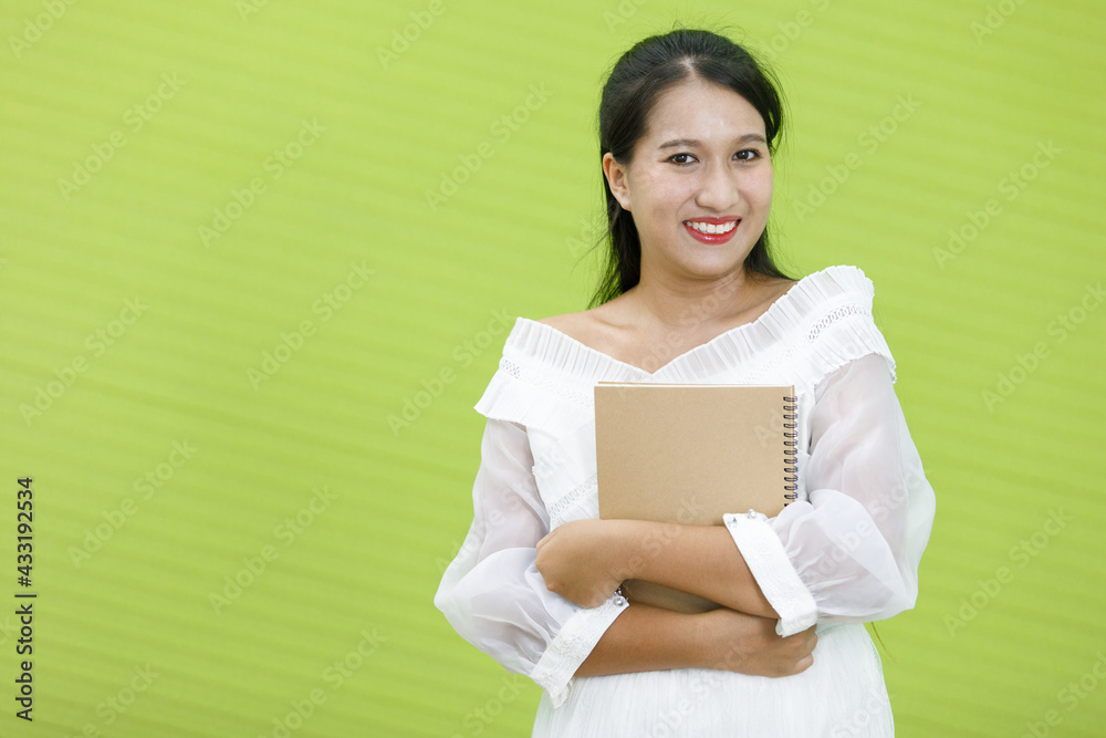 Image is portrait. Asian smiling plump woman wearing white dress. Lady asia standing and holding hand book in green background. Image is portrait. Asian smiling plump woman wearing white dress. Lady asia standing and holding hand book in green background.