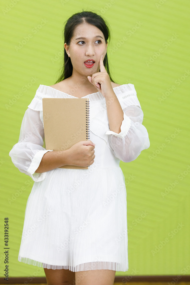 Image is portrait. Asian smiling plump woman wearing white dress. Lady asia standing finger poked on the cheek and holding hand book in green background. Image is portrait. Asian smiling plump woman wearing white dress. Lady asia standing finger poked on the cheek and holding hand book in green background.