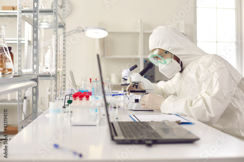 Unrecognizable scientist pharmacologist in disposable protective uniform using microscope working on medicine  biotechnology research in advanced pharma lab. Medical development laboratory