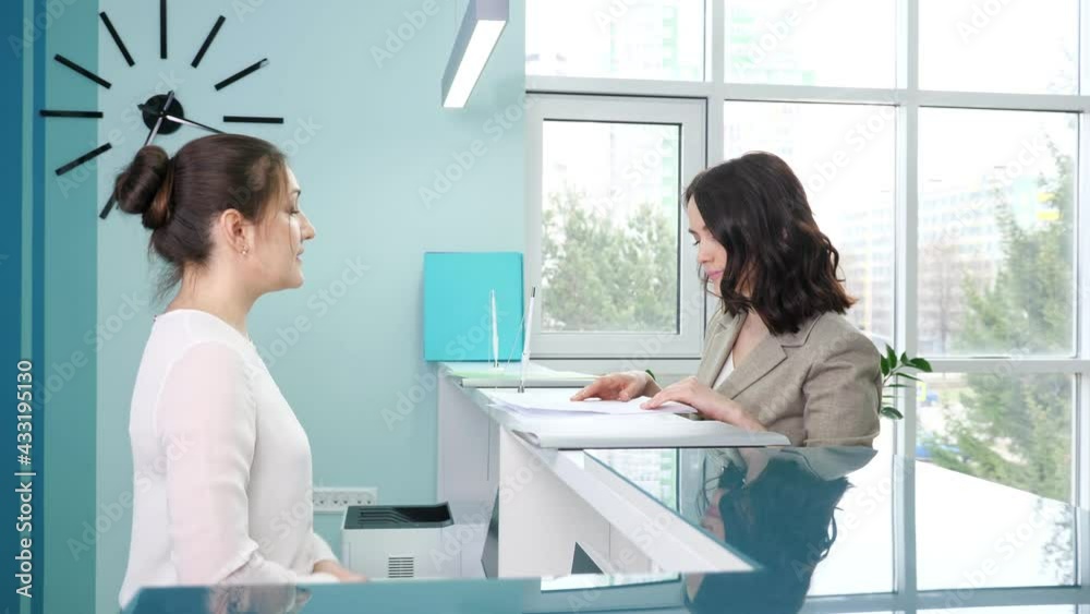 Woman receptionist with hair bun gives forms to fill in to lady client standing at reception counter in contemporary hospital close side view
