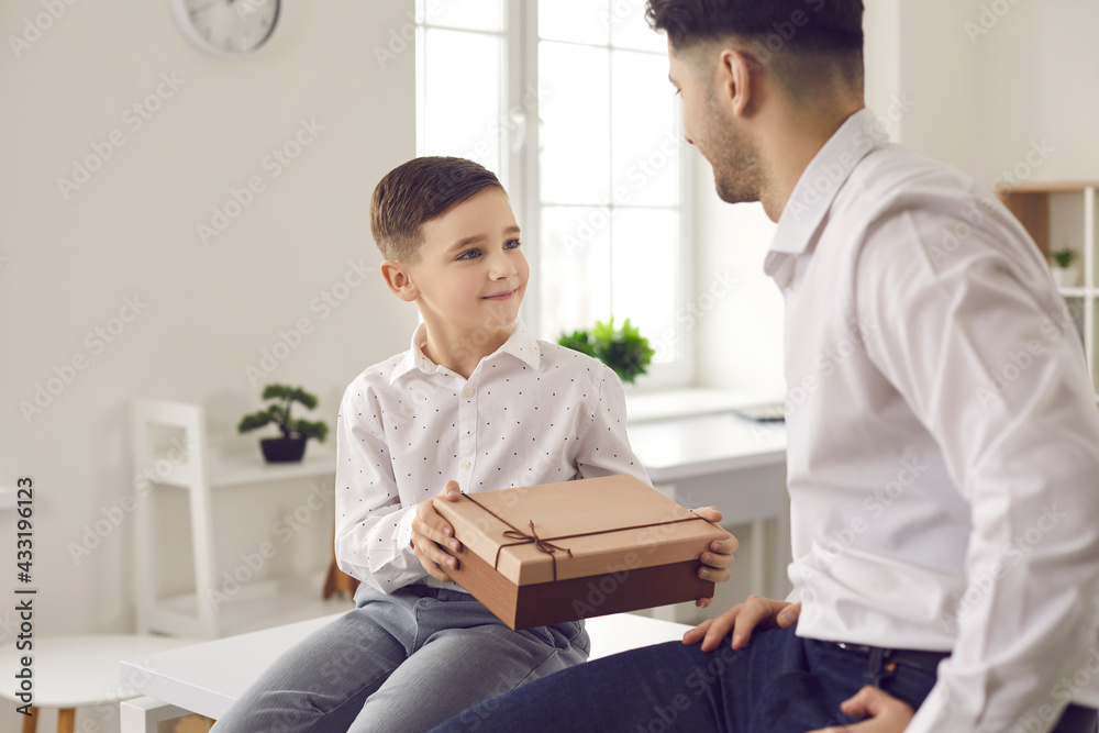 Child makes a pleasant surprise to the father. Cute little boy son gives dad a gift in a gift box in honor of his father's day or birthday. Concept of family celebration of important dates. Child makes a pleasant surprise to the father. Cute little boy son gives dad a gift in a gift box in honor of his father's day or birthday. Concept of family celebration of important dates.
