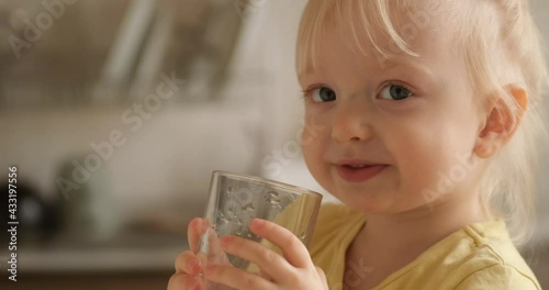 A little blonde girl drinks water from a glass in the kitchen looking at the camera and smiling