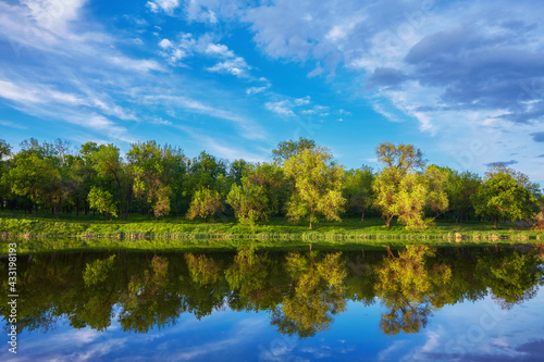 Beautiful river coast at sunset in summer. Colorful landscape with lake, green trees and grass, blue sky with multicolored clouds and orange sunlight reflected in water.