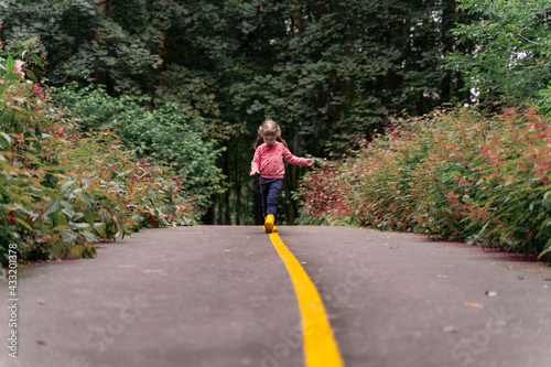 little girl in yellow rubber boots walks along the road in the park on a summer day