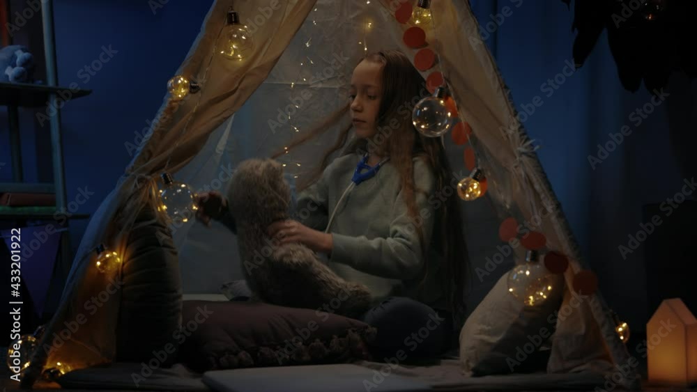 Cute girl wearing medical mask to teddy bear while sitting on floor in decorative makeshift tent at home. Kid with toy stethoscope playing in evening. Concept of leisure. Cute girl wearing medical mask to teddy bear while sitting on floor in decorative makeshift tent at home. Kid with toy stethoscope playing in evening. Concept of leisure.