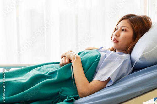 Portrait shot of Asian young hopeless upset coronavirus patient in blue hospital uniform lay down hold hands together with nomal saline solution drip tube injected look at camera on bed in ward room