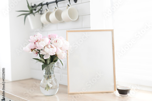 Vertical frame mockup on a wooden table in the kitchen. Glass vase with a bouquet of pink peonies and a cup of black coffee. Scandinavian style interior.