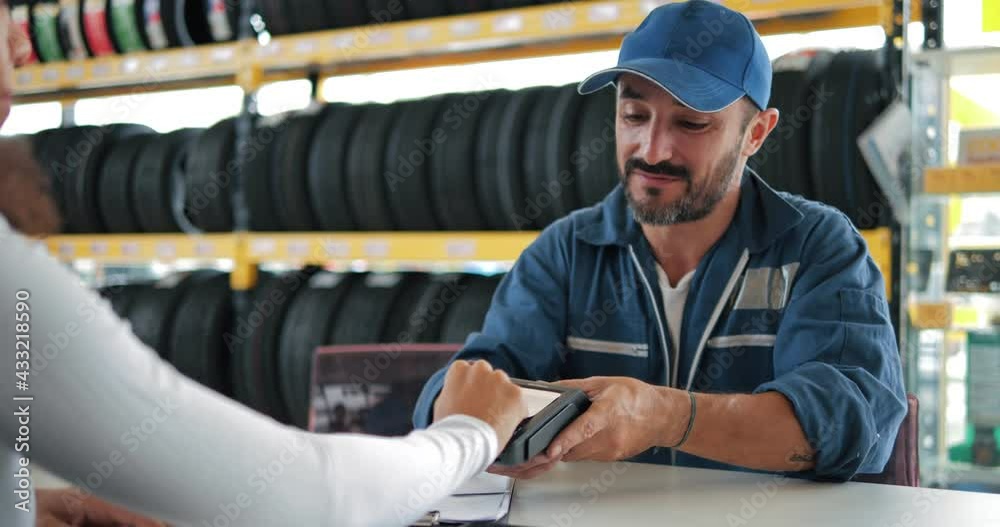 Customer paying with contactless credit card with NFC technology to worker of car tires service shop, credit card reader machine. Tires showing for sell or fix in the shop