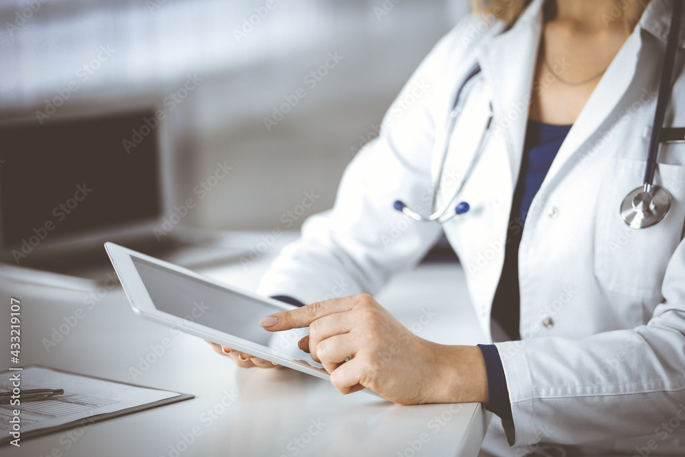 Unknown young woman-doctor is checking some medication names, while sitting at the desk in her cabinet in a clinic. Physician with a stethoscope is using a tablet computer, close-up. Perfect medical Unknown young woman-doctor is checking some medication names, while sitting at the desk in her cabinet in a clinic. Physician with a stethoscope is using a tablet computer, close-up. Perfect medical