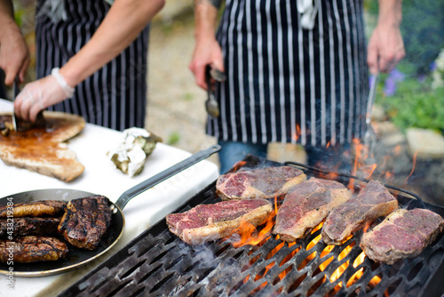 Steaks grilling on open flame and chef hands slicing steak in background