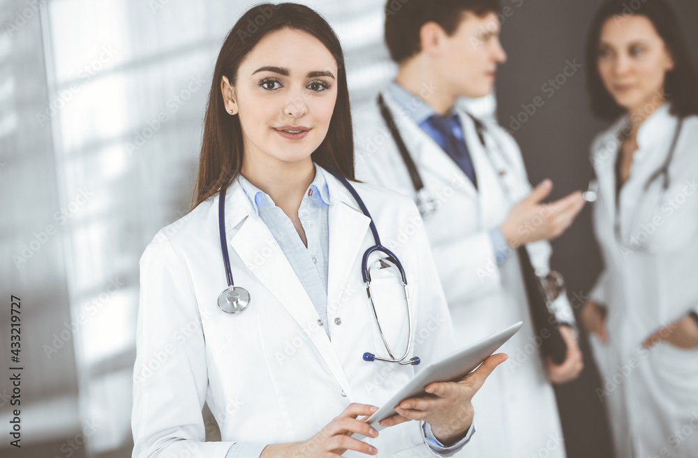 Smiling intelligent woman-doctor is holding a tablet computer in her hands, while she is standing together with her colleagues in a clinic. Physicians at work. Perfect medical service in a hospital Smiling intelligent woman-doctor is holding a tablet computer in her hands, while she is standing together with her colleagues in a clinic. Physicians at work. Perfect medical service in a hospital