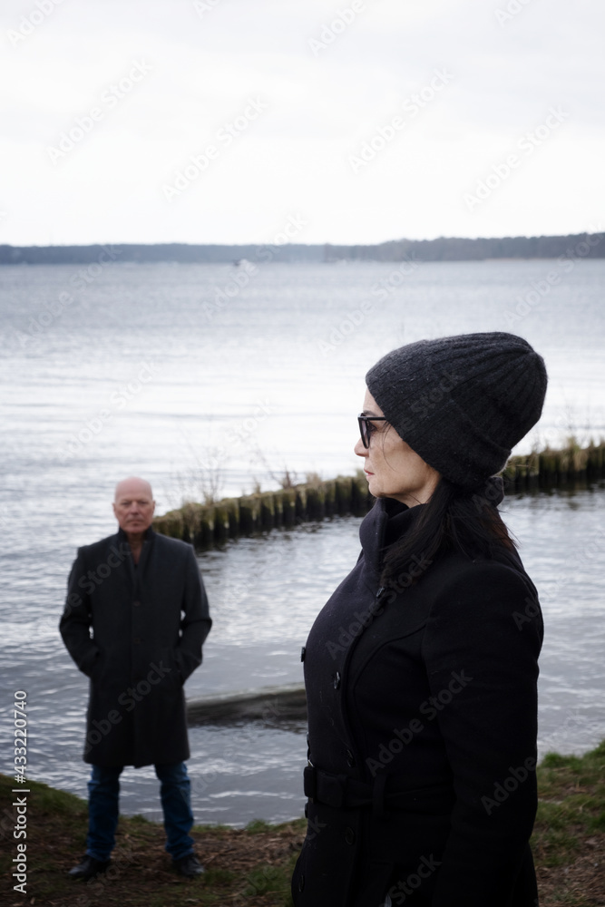 woman in black coat  standing at lake with man standing behind her in the distance