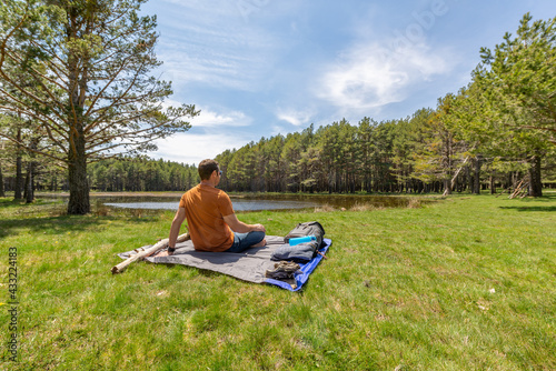 Back view of a traveler man sitting on a blanket close to a lake  located in the middle of a pine forest. Travel lifestyle concept. 