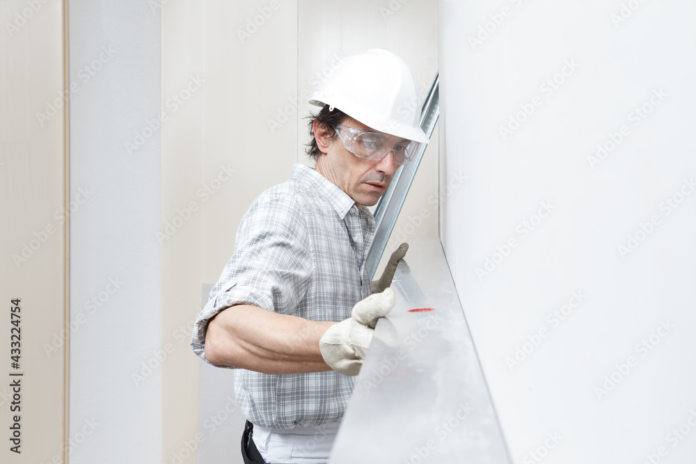 Man drywall worker or plasterer checking level of white plasterboard wall with bubble level at construction site. Wearing white hardhat, work gloves and safety glasses. Man drywall worker or plasterer checking level of white plasterboard wall with bubble level at construction site. Wearing white hardhat, work gloves and safety glasses.