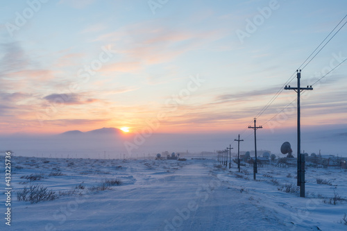 Road, electric poles and large satellite dishes in the snowy winter tundra. Industrial winter arctic landscape. Cold frosty weather. Blowing snow. Sunset. Chukotka, Siberia, Far East of Russia, Arctic