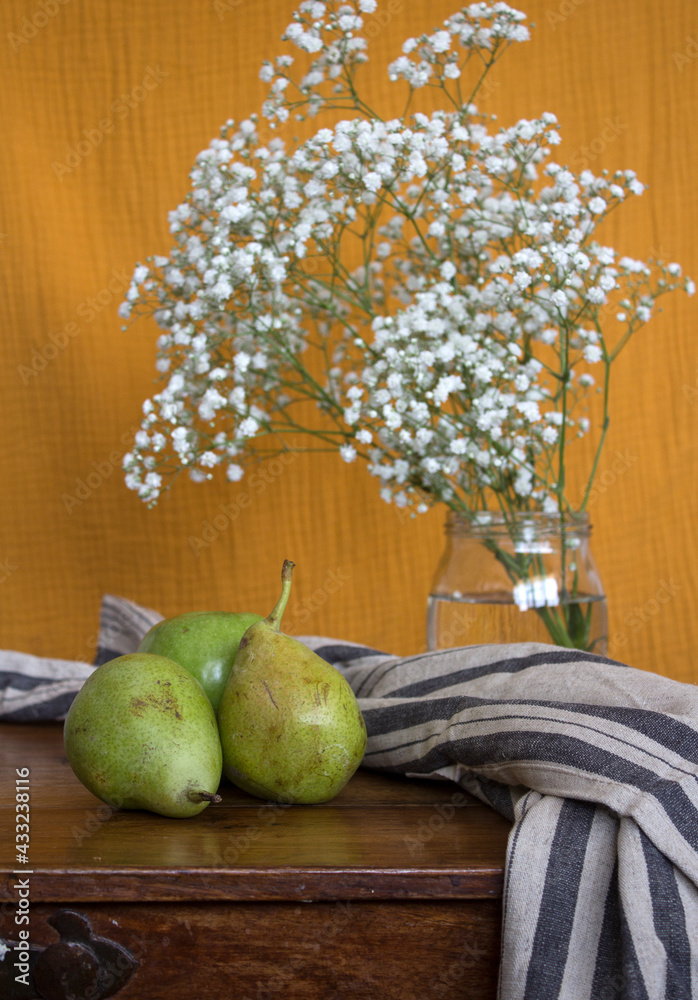 Still life with three pears. Glass jar with gypsophila flowers, linen kitchen table and green pears on wooden table. Summer still life with fresh fruits. Yellow textured background. 
