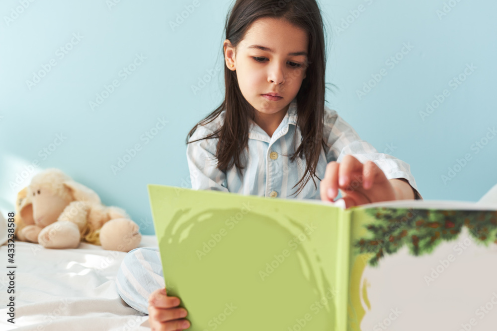 Little girl sitting in the bed and reading the fairy tale book. Kid in pajamas resting and reading interesting stories in a comfortable bed in the morning at home. Little girl sitting in the bed and reading the fairy tale book. Kid in pajamas resting and reading interesting stories in a comfortable bed in the morning at home.