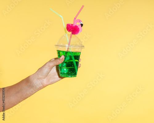 Close up shot of female hand holding refreshing tropical cocktail drink with flamingo straw isolated on yellow background