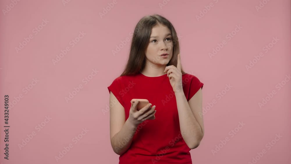 Portrait of fashion model smiling and texting on her phone. Young girl with long hair in red t-shirt posing on pink studio background. Close up. Slow motion ready 59.94fps. Portrait of fashion model smiling and texting on her phone. Young girl with long hair in red t-shirt posing on pink studio background. Close up. Slow motion ready 59.94fps.