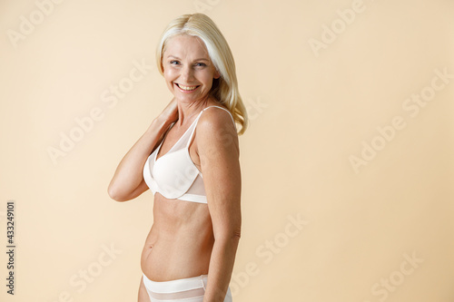 Portrait of happy mature woman with blonde hair in white underwear smiling at camera, posing isolated over beige background