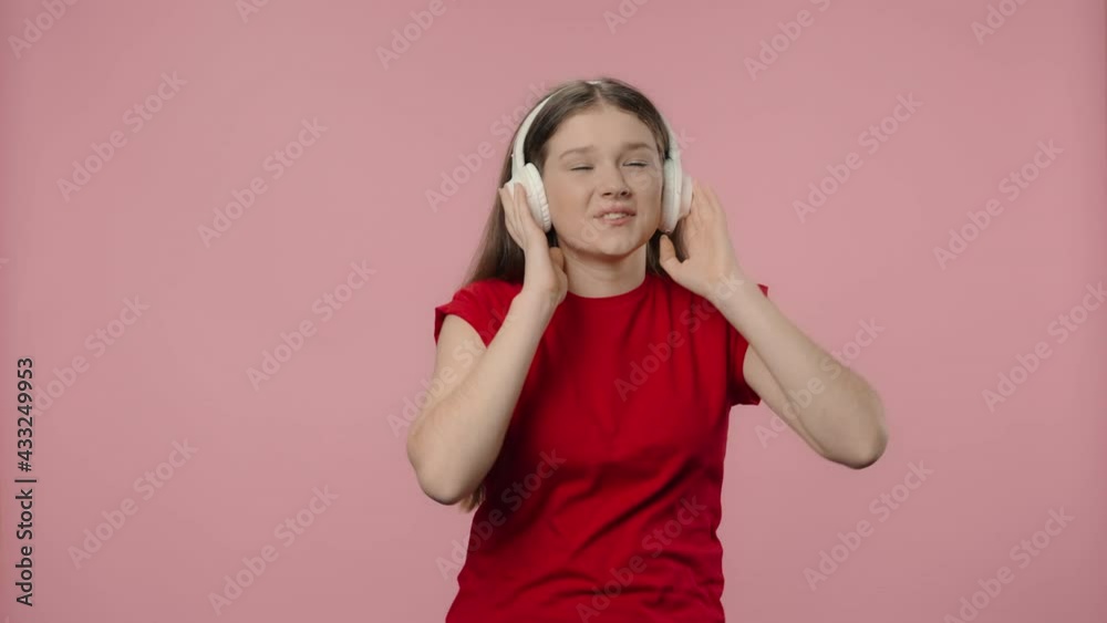Portrait of fashion model enjoying music in big wireless white headphones. Young girl with long hair in red t-shirt posing on pink studio background. Close up. Slow motion ready 59.94fps. Portrait of fashion model enjoying music in big wireless white headphones. Young girl with long hair in red t-shirt posing on pink studio background. Close up. Slow motion ready 59.94fps.