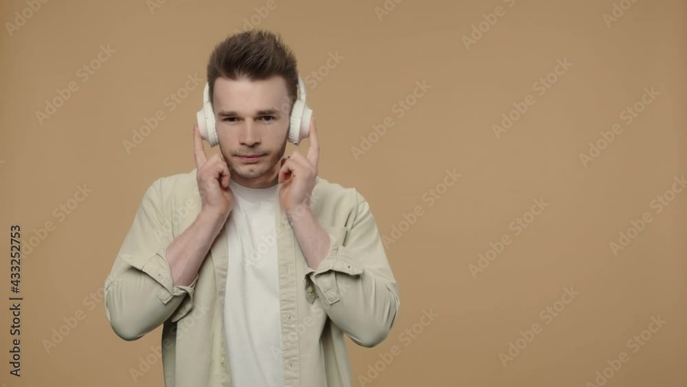 Portrait of stylish guy enjoying music in big wireless white headphones. Male fashion model in a beige shirt posing on brown studio background. Close up. Slow motion ready 59.94fps. Portrait of stylish guy enjoying music in big wireless white headphones. Male fashion model in a beige shirt posing on brown studio background. Close up. Slow motion ready 59.94fps.