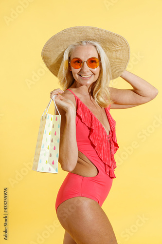 Studio shot of stylish mature blonde woman in red swimsuit and straw hat smiling at camera while posing with shopping bag, standing isolated on yellow background