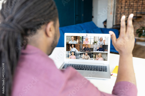 African-American man with locks hairstyle using laptop computer for virtual meeting with diverse colleagues or friends, waving hello, back view at the monitor with group of colleagues on it