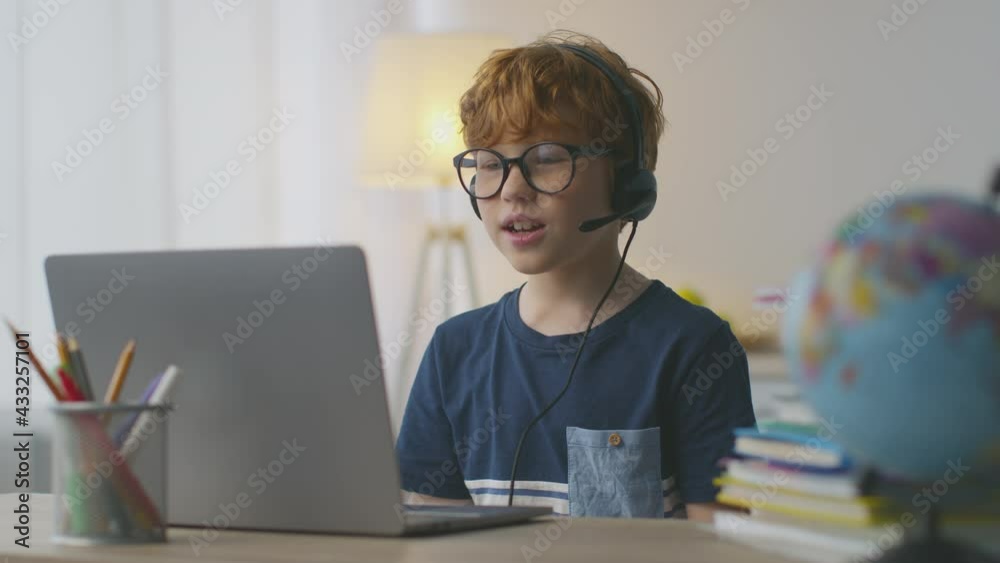 Little redhead boy wearing headset with mic video chatting with tutor, looking at laptop and talking to teacher