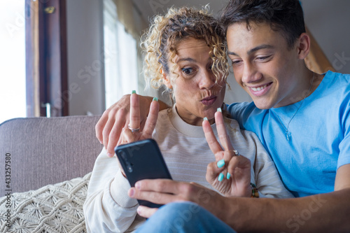 Smiling middle-aged 40s mother rest with grown-up son using smartphone together, happy young man enjoy family weekend with mom browsing wireless Internet on cellphone, have fun at home
