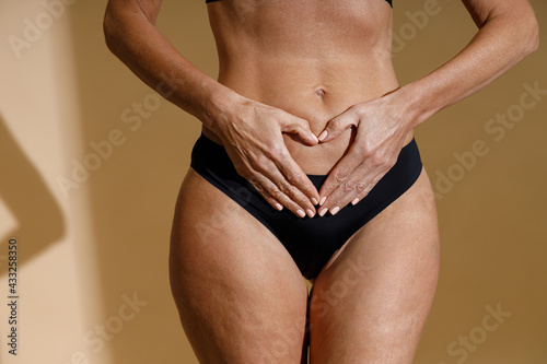 Close up shot of woman's hands forming a heart symbol on belly. Perfect fit body of mature woman in black underwear holding hands on her stomach while standing isolated over beige background