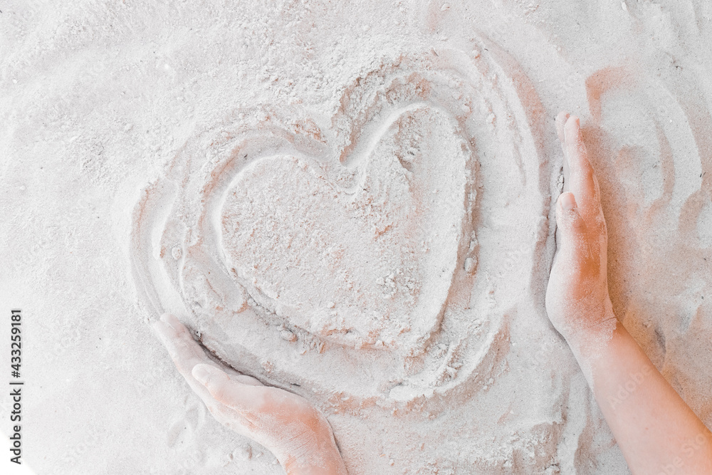 Obraz premium The hand of a young girl draws a heart on the white beach sand close-up. Symbol or sign of love concept