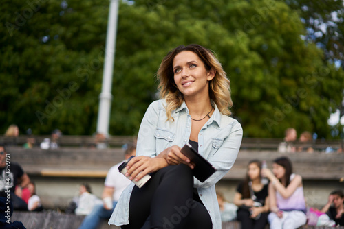 Happy hipster girl in casual clothes using modern touch pad during resting time in city enjoying leisure, cheerful female blogger with digital tablet holding ebook and takeaway coffee to go