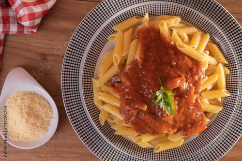 Macarrão pene com molho de tomate acompanhado de queijo ralado. Fundo em madeira com toalha xadrez dobrada.