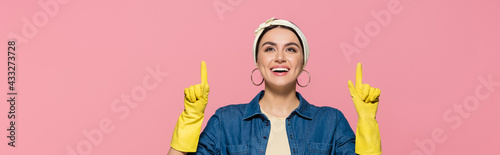 Smiling housewife in yellow rubber gloves pointing up with fingers isolated on pink, banner