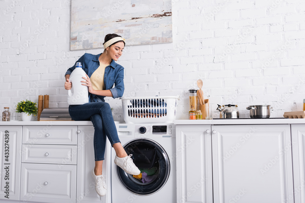 Cheerful housewife with detergent sitting near washing machine and clothes Cheerful housewife with detergent sitting near washing machine and clothes