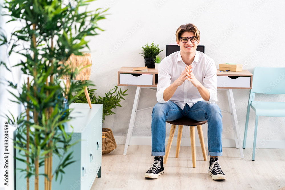 Smiling young man sitting with hands clasped on stool in living room Smiling young man sitting with hands clasped on stool in living room