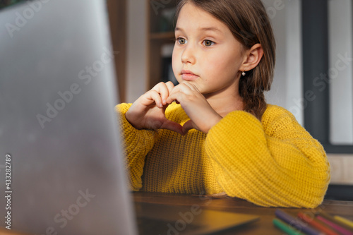 Cute little girl making heart shape with hand during video call on laptop at home photo