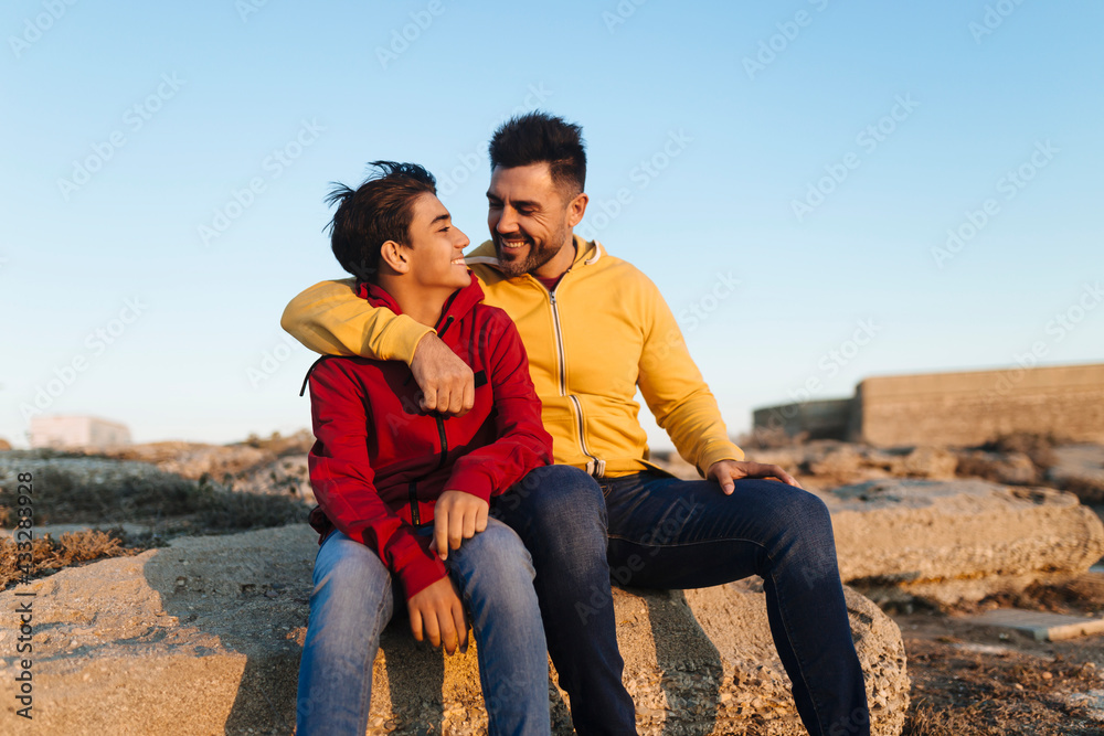 Happy father and son sitting on rock against sky during sunset Happy father and son sitting on rock against sky during sunset