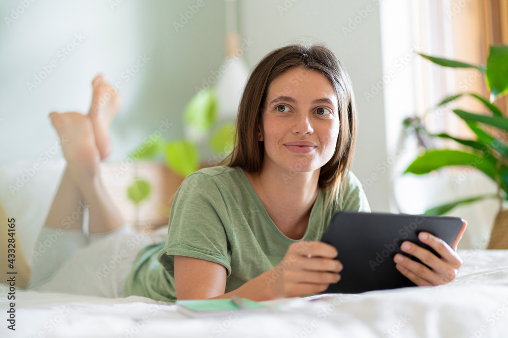 Woman with digital tablet looking away while lying on bed at home Woman with digital tablet looking away while lying on bed at home