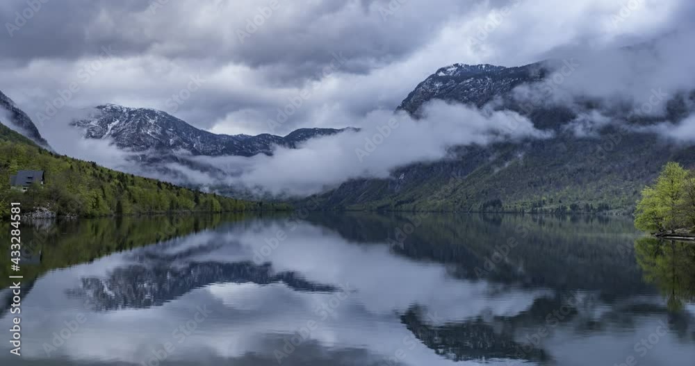Time lapse of clouds moving over Alps mountains and Alpine lake Bohinj in Slovenia. Dramatic cloudy day. Reflection in pristine lake. Wide angle, right trucking