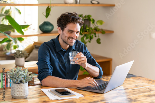 Smiling male freelance worker holding glass of water while working on laptop at home office photo