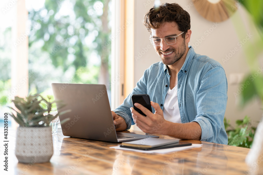 © steve brookland/Westend61 - Smiling male freelancer with eyeglasses using smart phone while sitting with laptop at home office