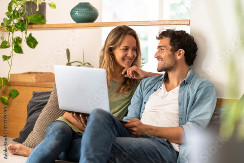 Smiling man with laptop looking at girlfriend while sitting on couch at home