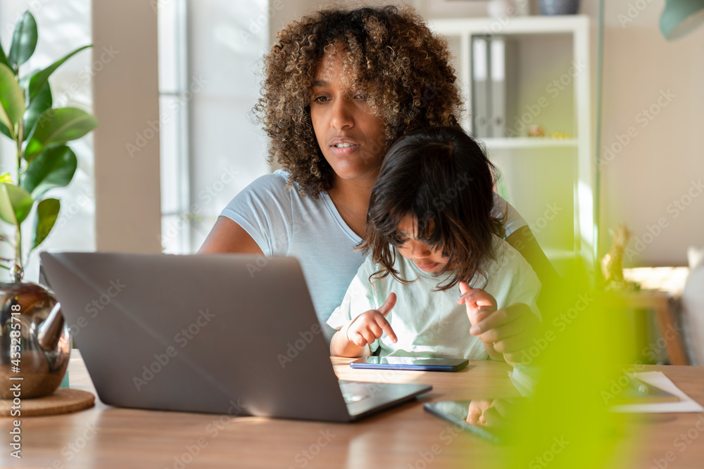Mother working on laptop while her daughter playing with smart phone in home office Mother working on laptop while her daughter playing with smart phone in home office