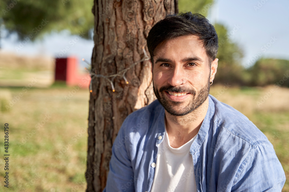 Smiling hipster man against tree trunk during sunny day Smiling hipster man against tree trunk during sunny day