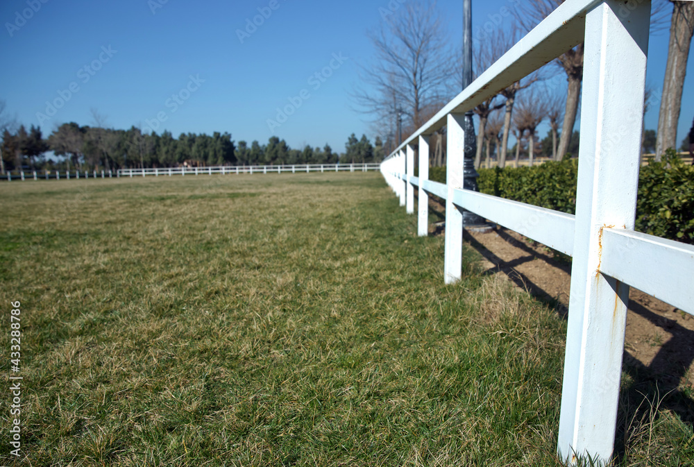 A white fence with green gardens . Horse racecourse field . Large farmland and ranch protection background . Green pastures of horse farms. Country summer landscape. White fence is a long way.