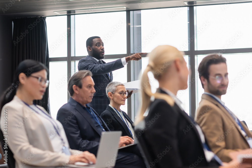 African american businessman pointing with finger near blurred colleagues in conference room African american businessman pointing with finger near blurred colleagues in conference room