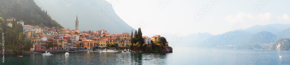 Fototapeta premium Early morning view of the Como Lake with the village of Varenna, Italy, and clear sky, with fog, soft sunlight and mountains in the distance